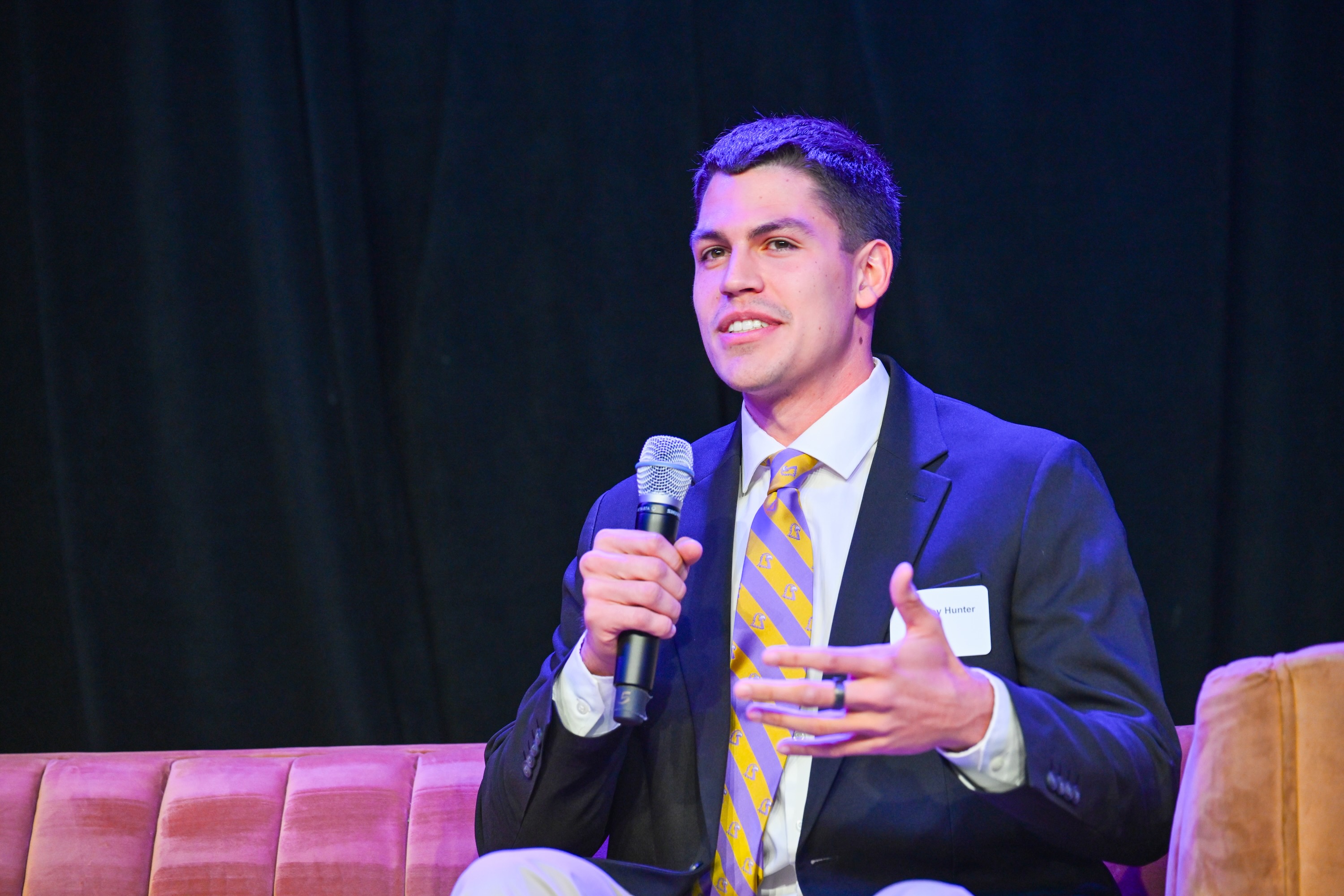 Westley Hunter speaking at a microphone in a dark suit at a panel event